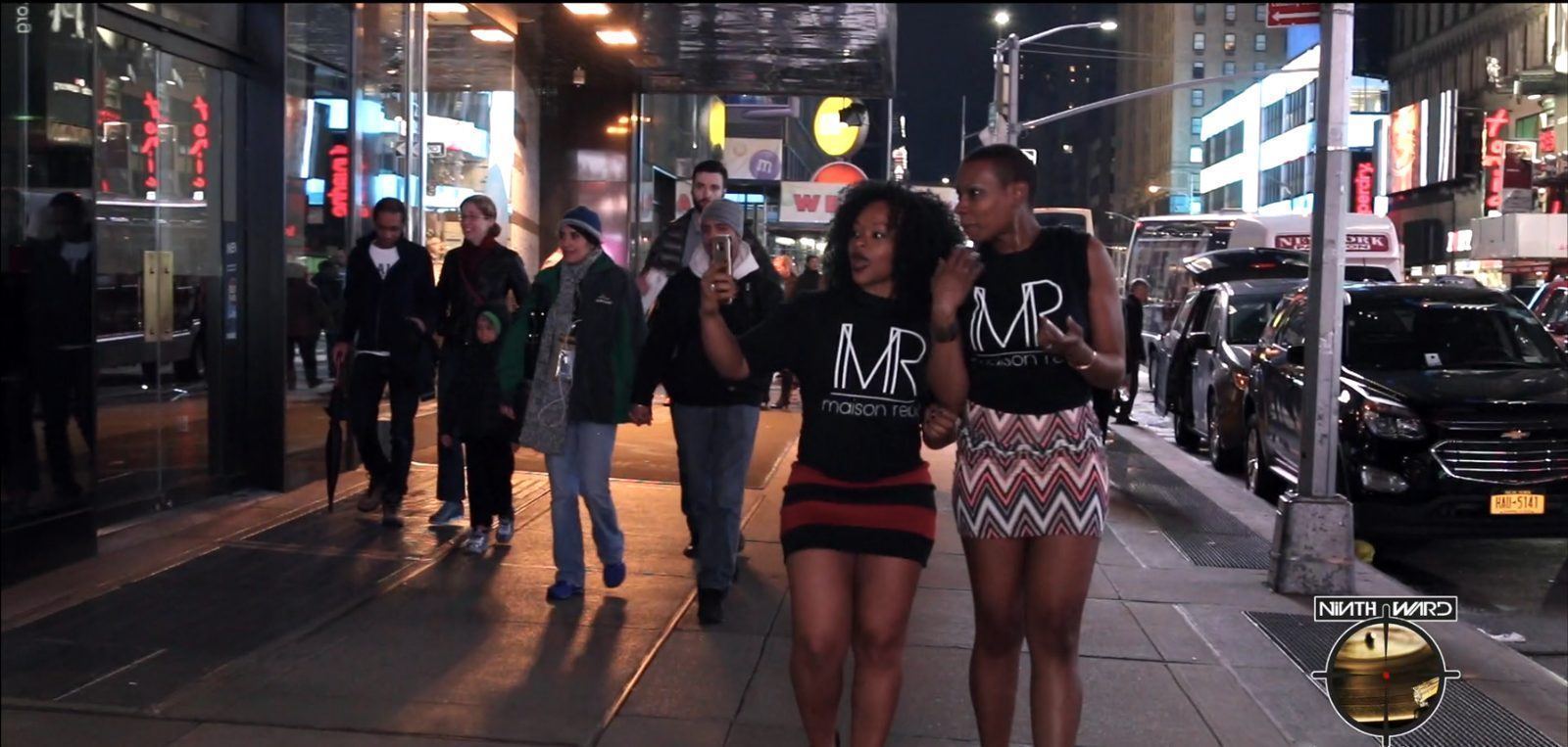 Events — Two brand ambassadors in matching MR shirts filming with a phone along a busy nighttime city sidewalk