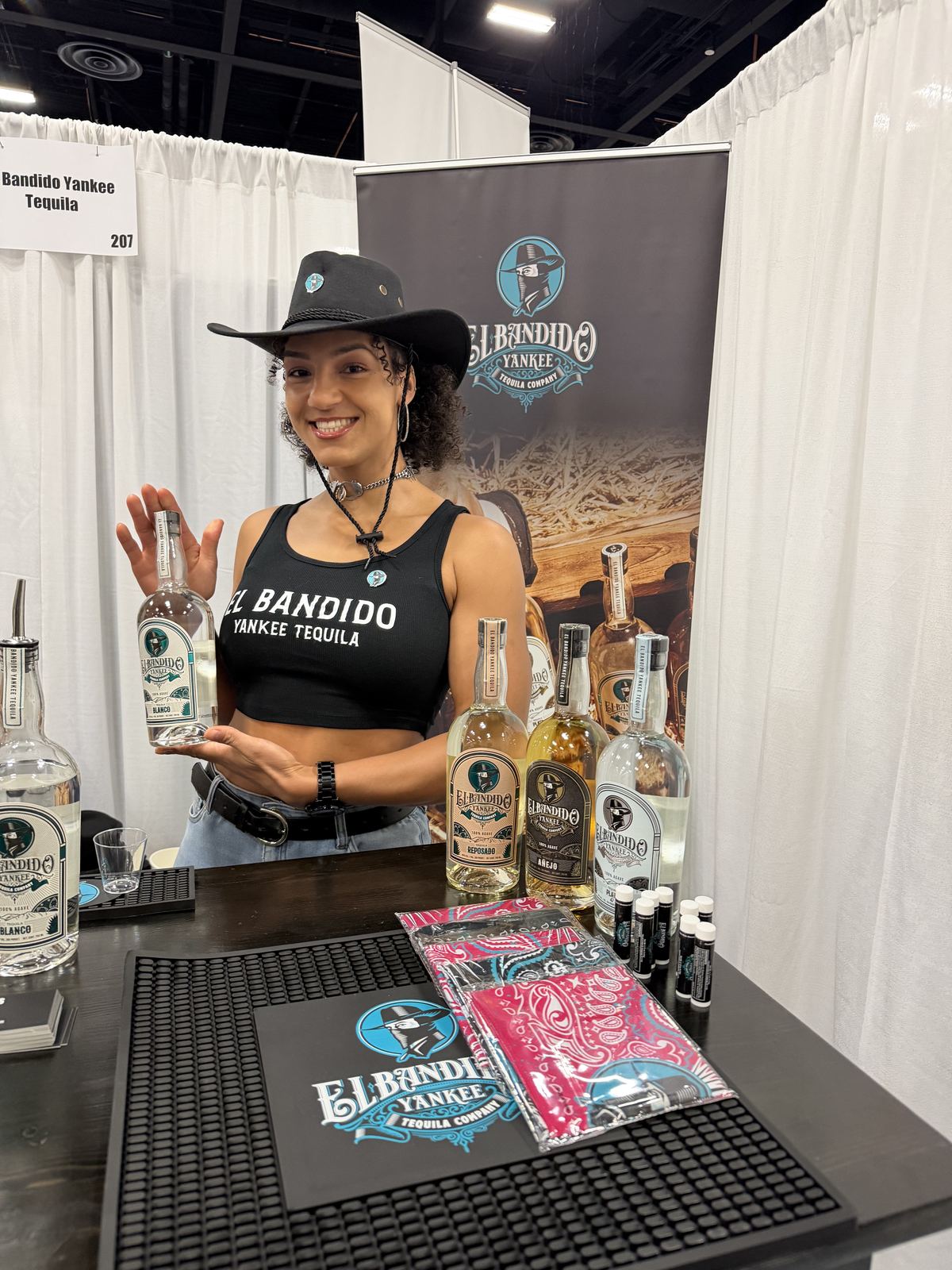 Events — Ambassador presenting El Bandido Yankee Tequila Blanco at booth 207 beside the full bottle lineup and branded bandanas