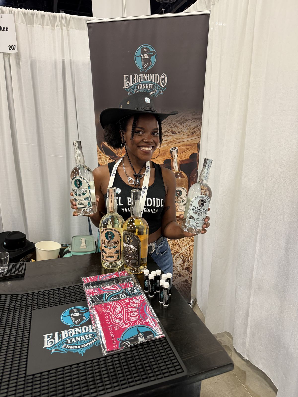 Events — Ambassador holding four El Bandido Yankee Tequila bottles behind the booth bar, Reposado and Añejo up front