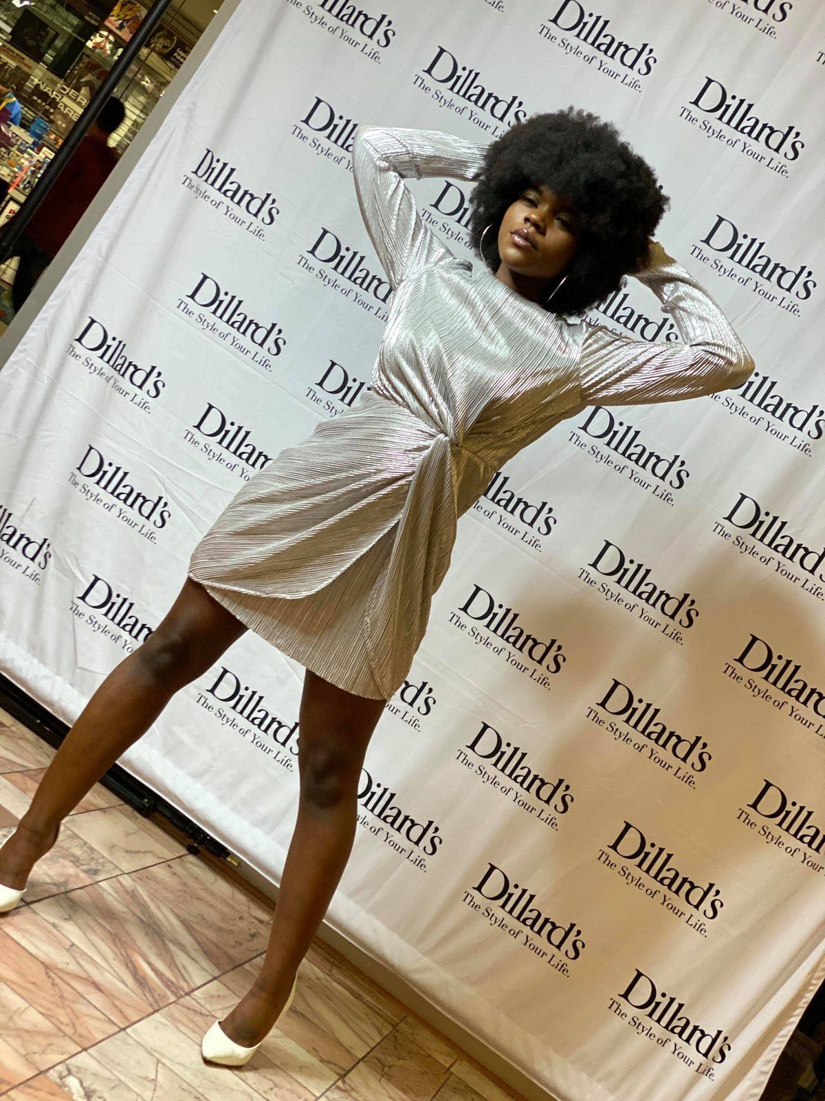Fashion Showcase — Model in a metallic silver wrap dress and hoop earrings posing at a Dillard's step-and-repeat backdrop