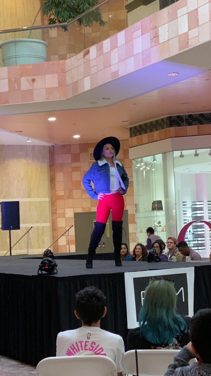 Fashion Showcase — Model in a wide-brim hat, sherpa denim jacket and thigh-high boots posing on a mall runway stage