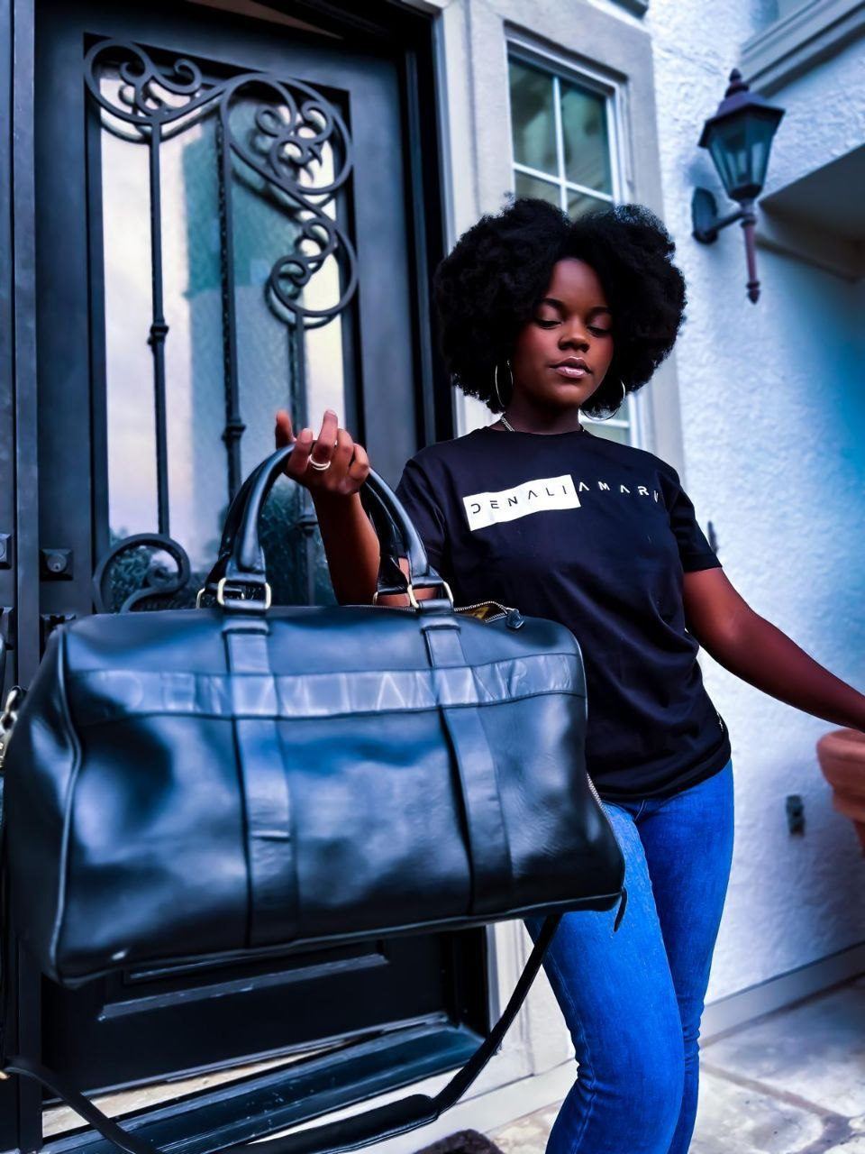 Influencer Work — Creator in a Denali Amaru logo tee holding a black leather duffel outside an ornate iron front door