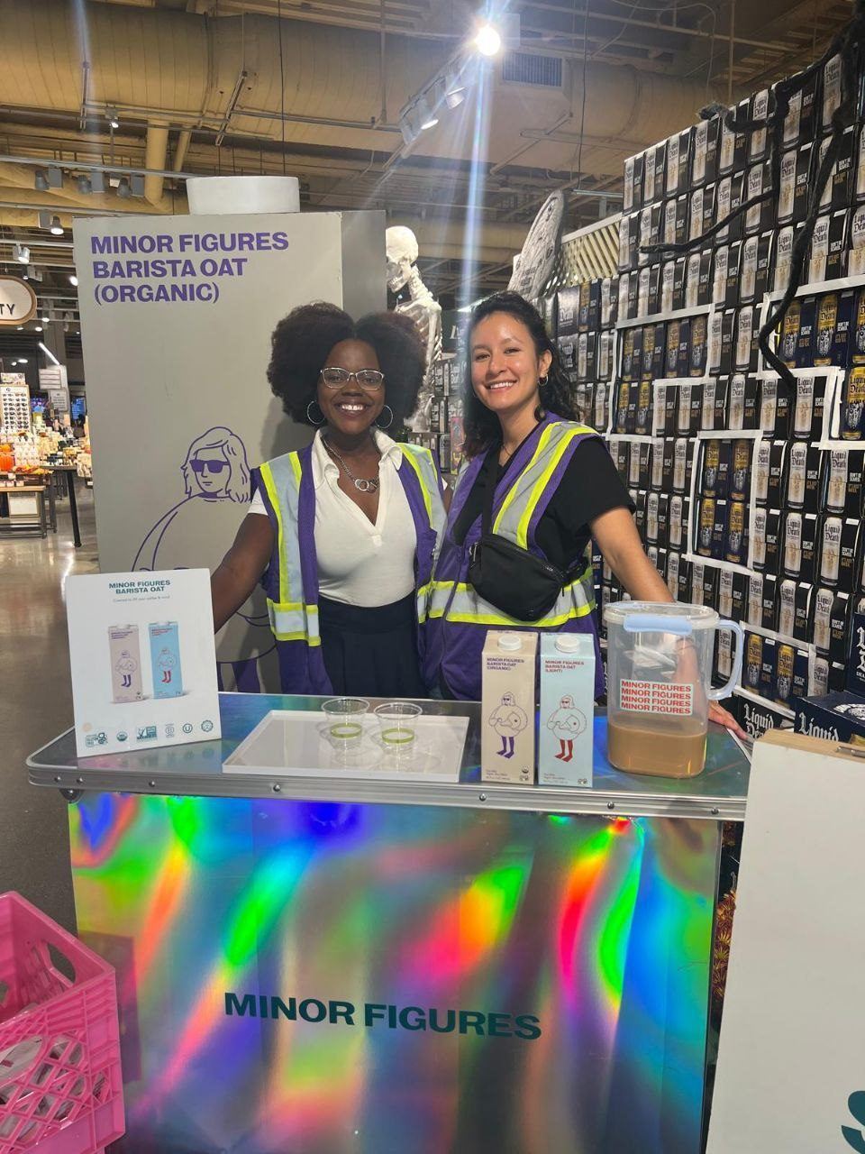 On-Site Activations — Two brand ambassadors sampling Minor Figures Barista Oat at a holographic demo cart beside stacked Liquid Death cases