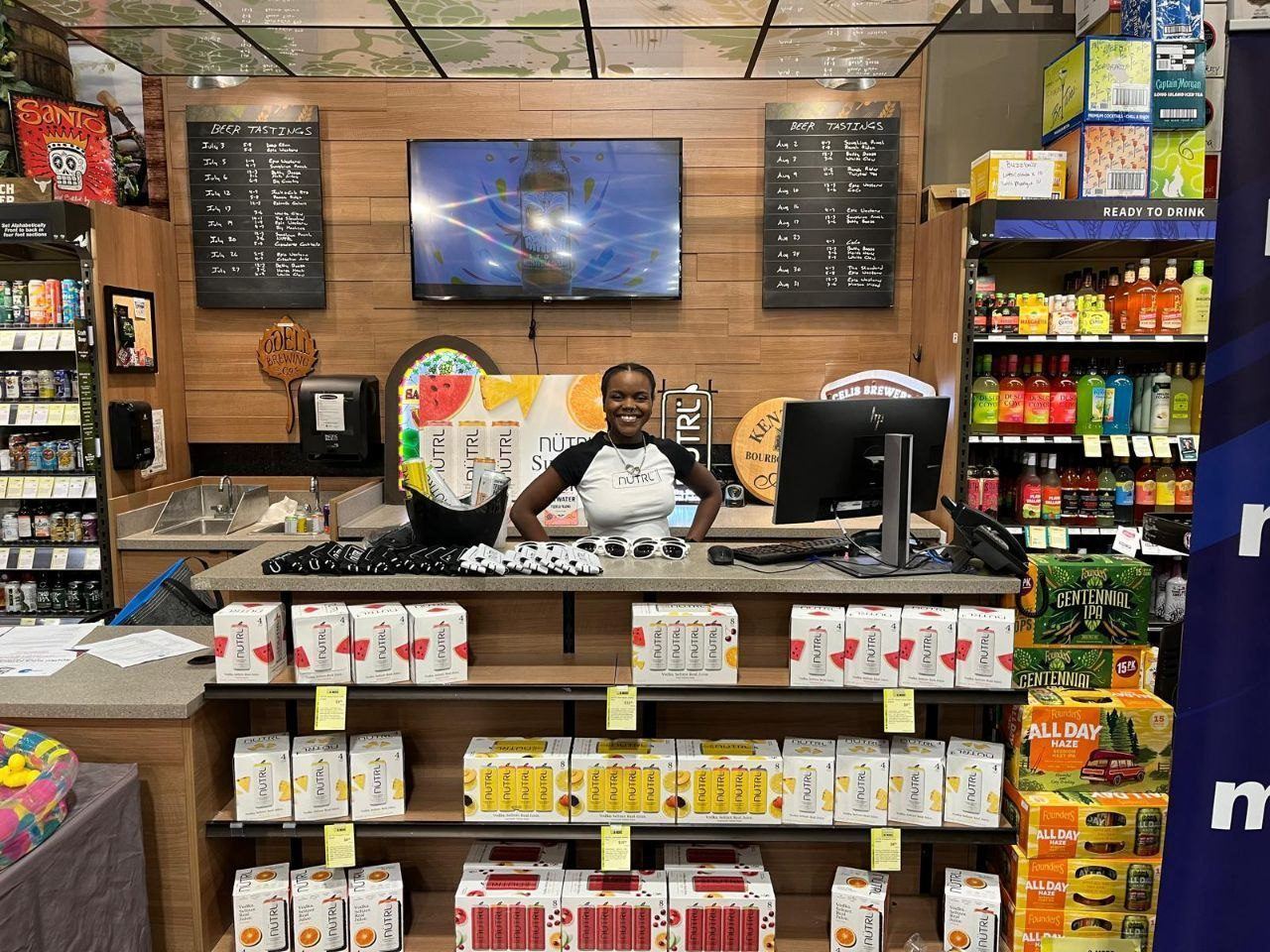 On-Site Activations — NUTRL ambassador behind a tasting counter stocked with vodka seltzer packs, branded sunglasses, and Founders IPA cases