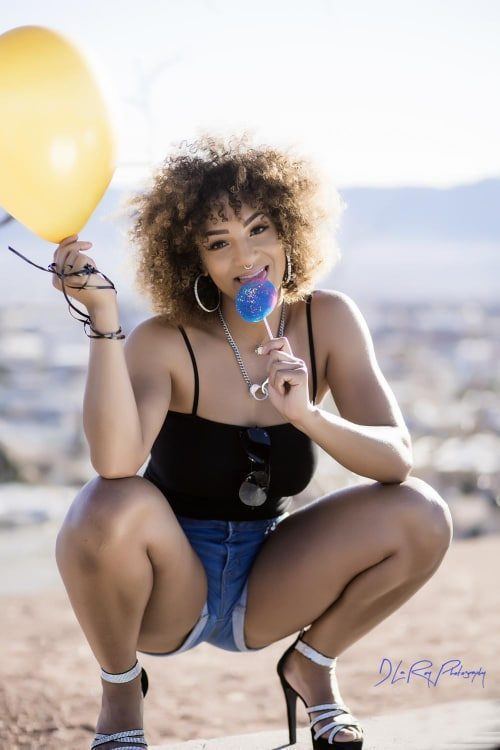 Photography — Model crouching on a hilltop overlook with a yellow balloon and swirl lollipop, hazy cityscape behind