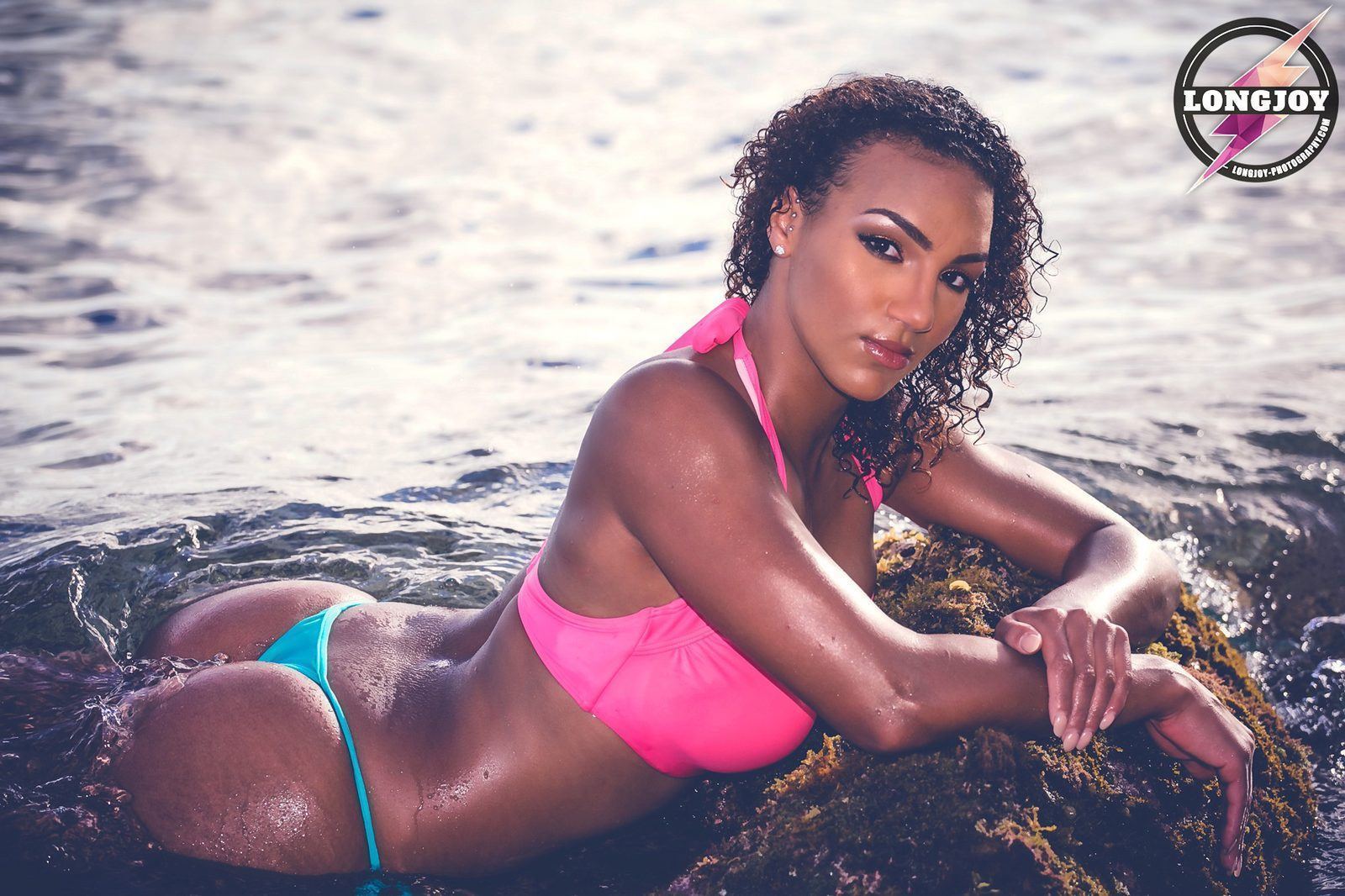 Photography — Swimwear model in a pink and teal bikini reclining against a mossy rock in ocean surf