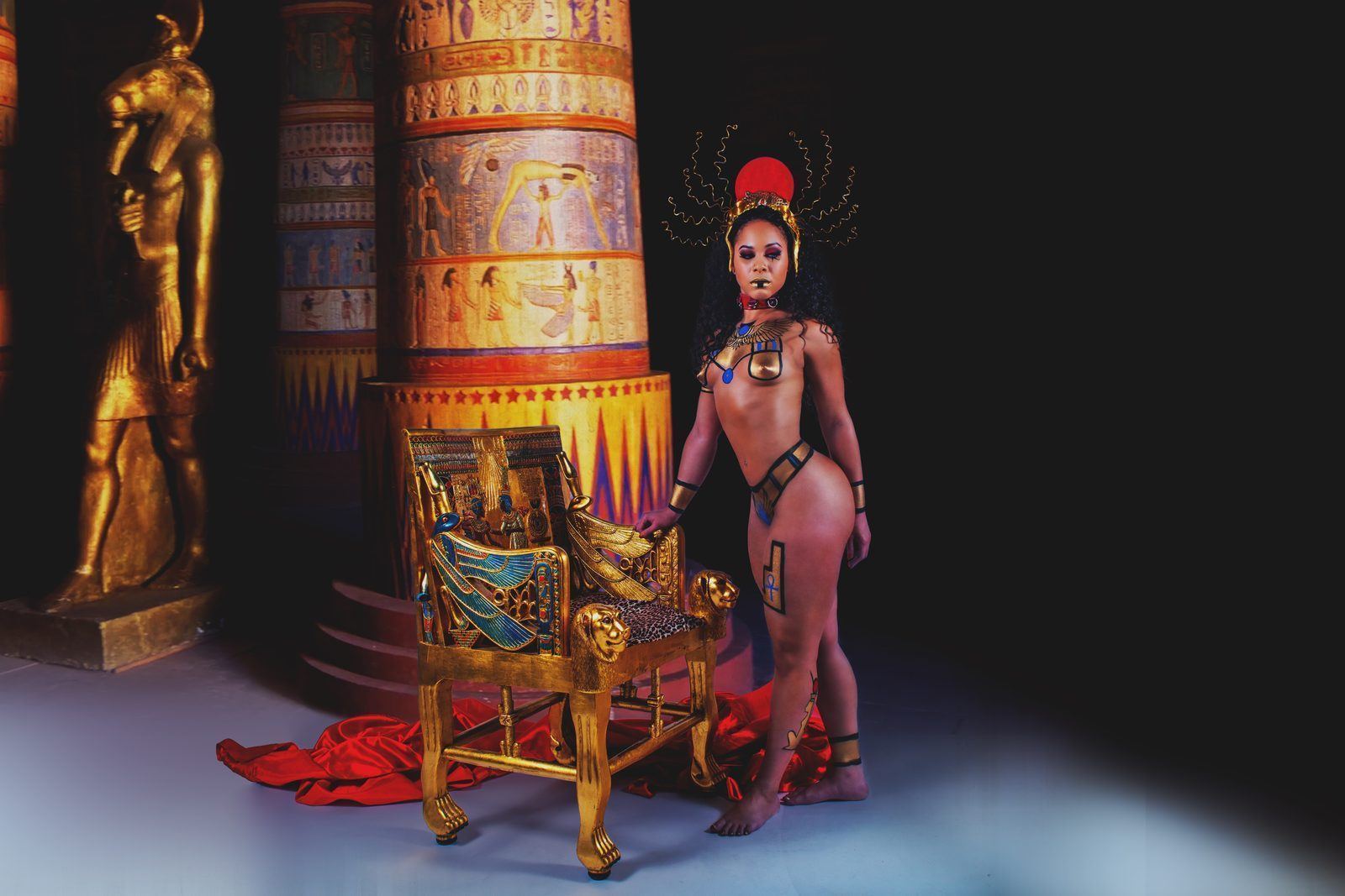Photography — Egyptian-themed editorial with a model in gold body paint and sunburst headdress beside a pharaoh throne and hieroglyphic columns