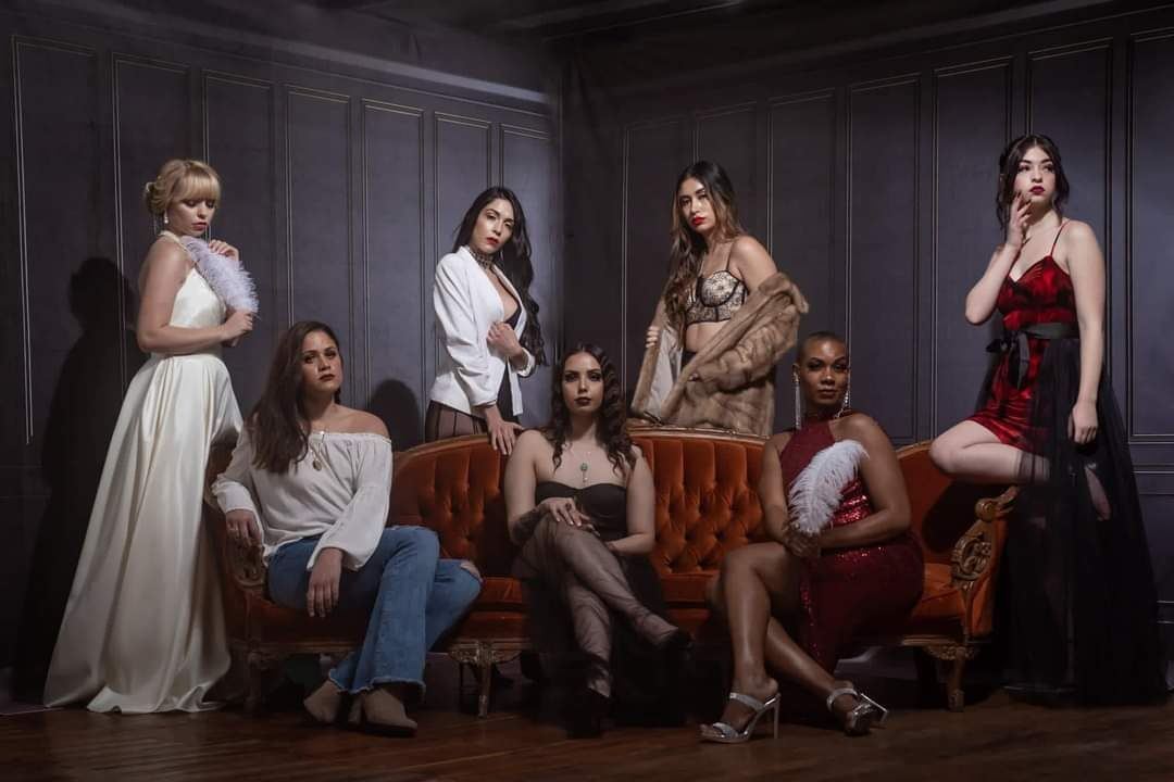 Photography — Seven models in evening and casual looks posed around a vintage tufted velvet sofa in a dark-paneled studio