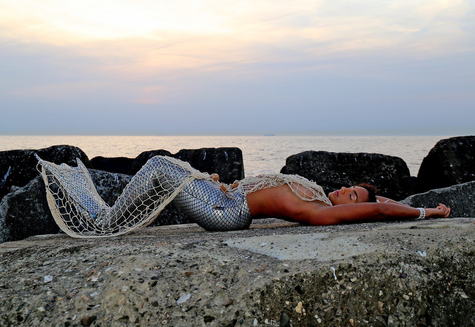 Photography — Model in a silver sequined mermaid tail draped with fishing net reclining on seaside rocks at sunset