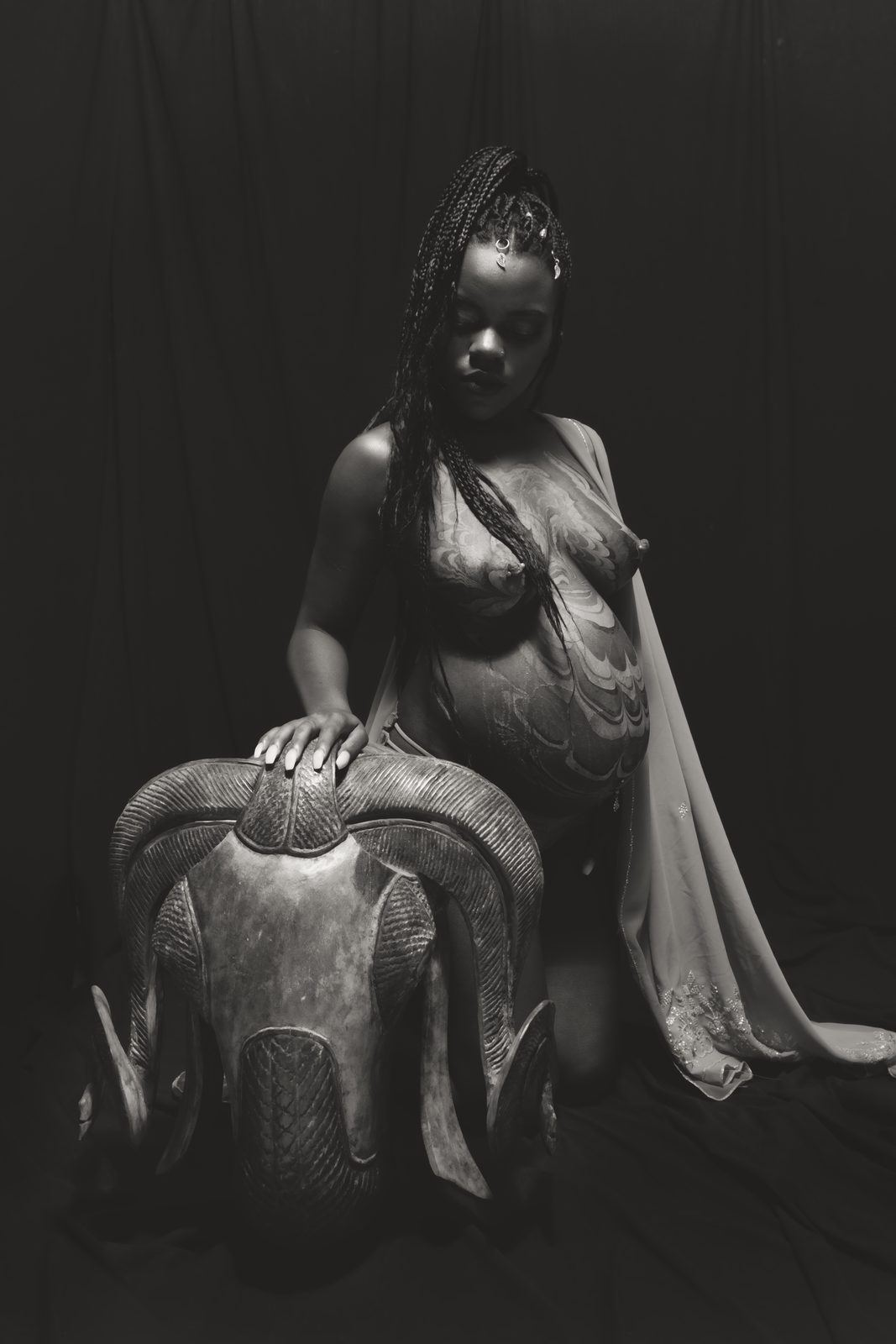 Photography — Black and white maternity portrait of a kneeling model with body paint resting a hand on a carved ram sculpture