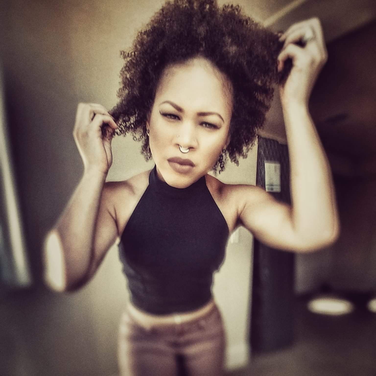 Photography — Model in a black halter crop top fluffing her curly hair for a close-up indoor portrait, septum ring visible