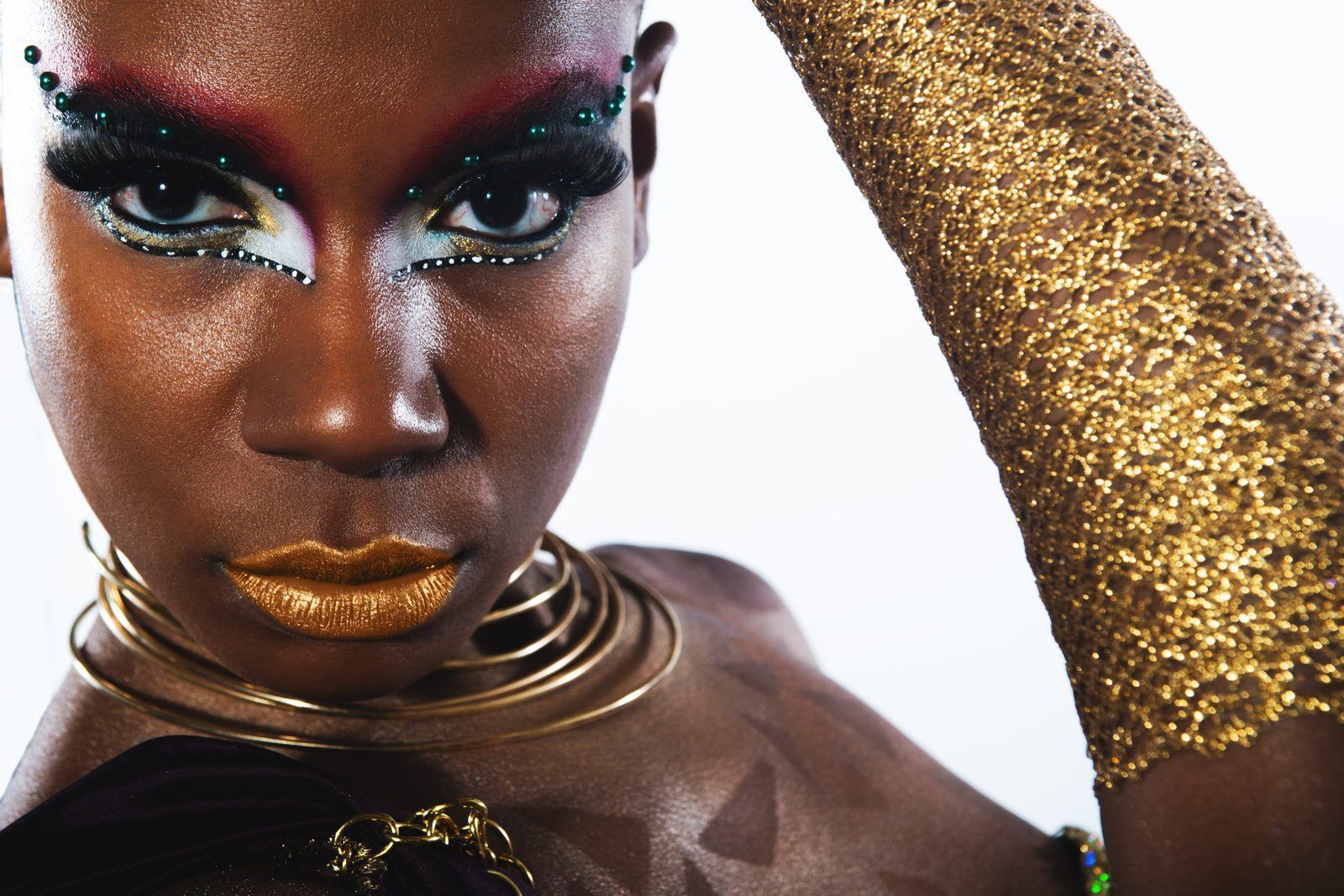 Photography — Close-up beauty portrait of a model with gold lips, jeweled red eyebrows, coiled gold choker, and glittering sleeve