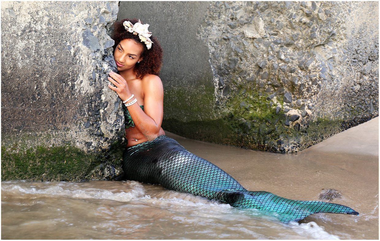 Photography — Model in an iridescent green mermaid tail and seashell crown posing against beach rocks in the surf