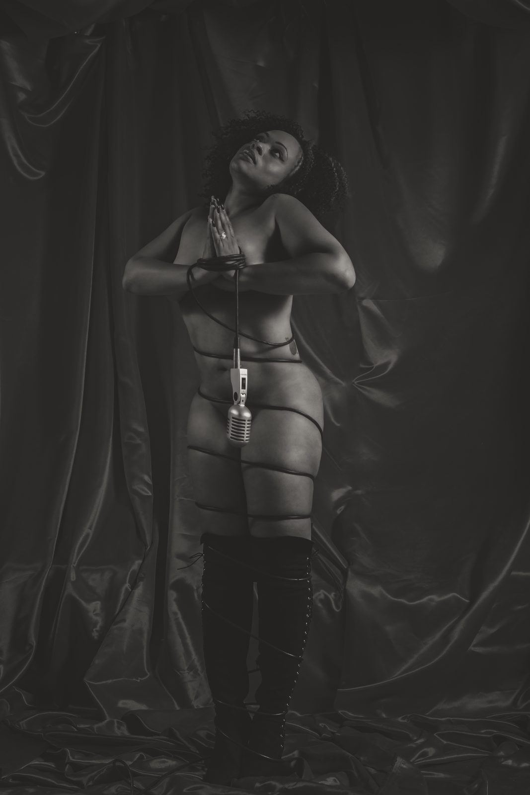 Photography — Model in thigh-high boots wrapped in a microphone cord, black-and-white studio portrait against satin drapery