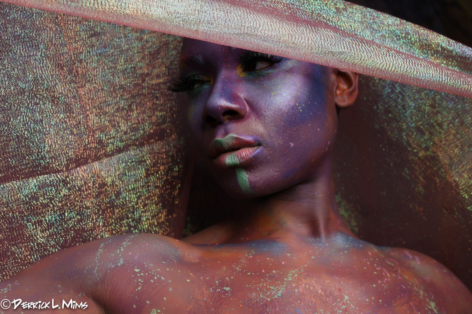 Photography — Close-up of a model in metallic body paint with paint speckles, framed by shimmering gold fabric