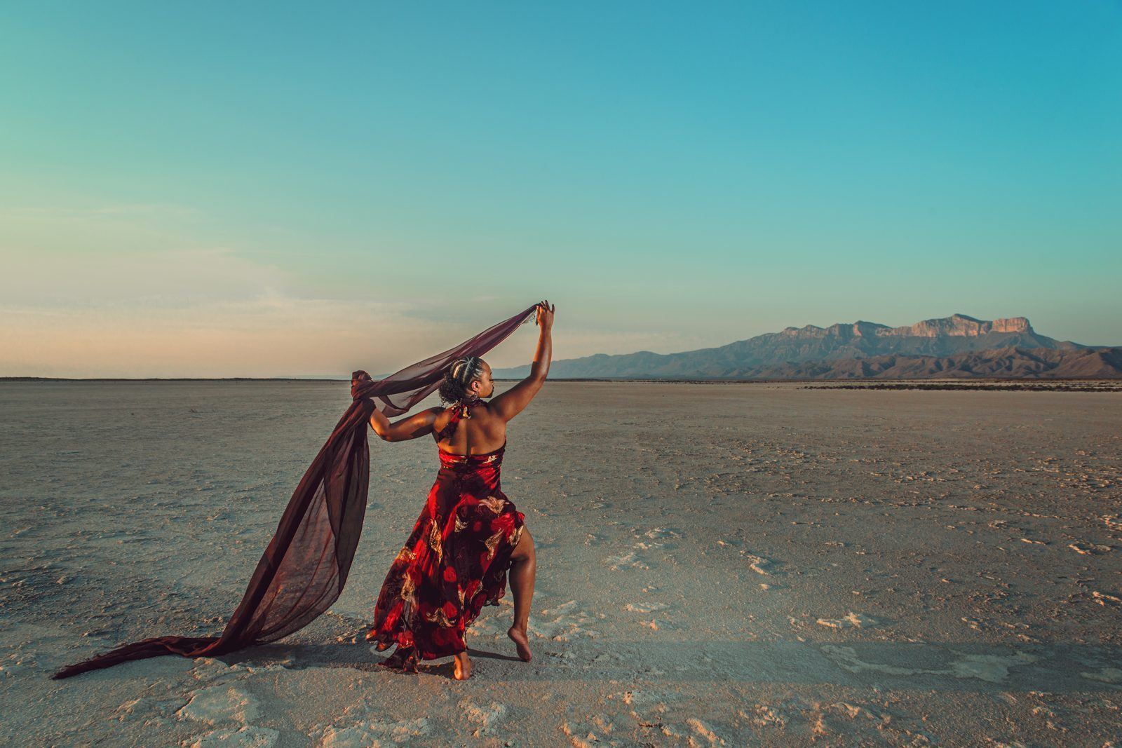 Photography — Model in a red print dress trailing a sheer scarf across a salt flat with mountains behind