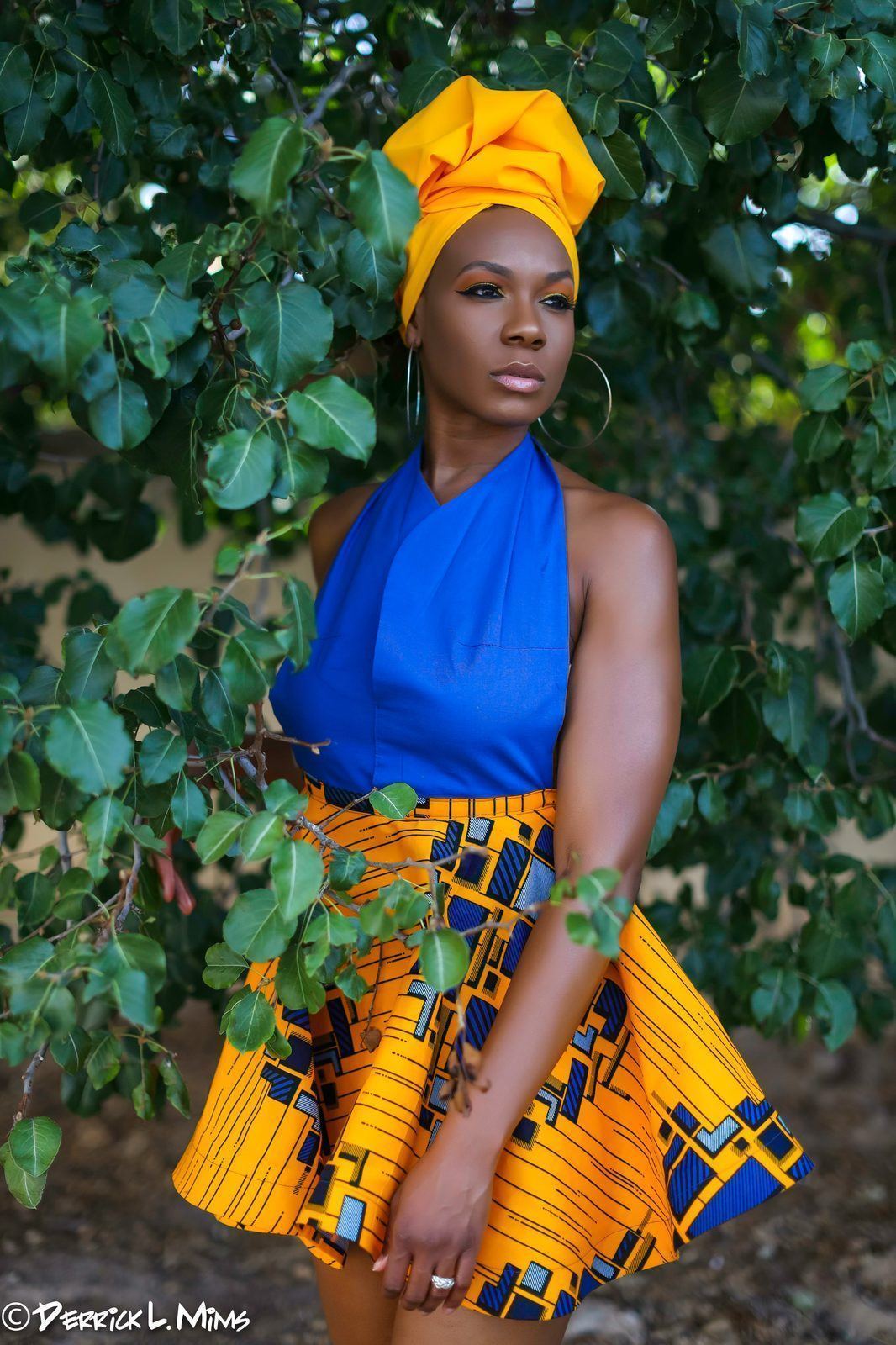 Photography — Model in a yellow head wrap, blue halter top, and geometric print skirt posing beneath leafy branches